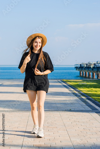 Young woman with long brown hair wearing a black outfit and straw hat walks along a waterfront promenade with blue water and pier in the background