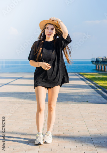 Young woman wearing a black oversized t-shirt and straw hat walks along a sunny waterfront promenade with blue water and clear sky in the background