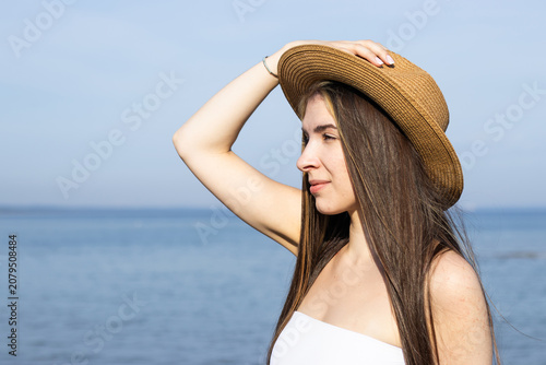 Young woman with long brown hair wearing a straw hat poses by the beach, with calm ocean waves and clear blue sky visible in the background