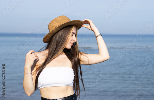 Young woman with long hair wearing a straw hat and white crop top poses by the ocean, with calm water and clear blue sky in the background