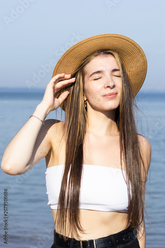 Young woman with long hair wearing a straw hat and white crop top stands by the beach, enjoying the sunlight with the ocean in the background