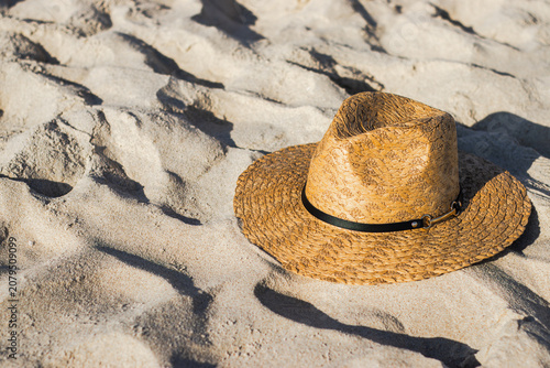 Straw sun hat resting on sandy beach surface with textured patterns and warm sunlight illuminating the scene, ideal for summer and vacation themes