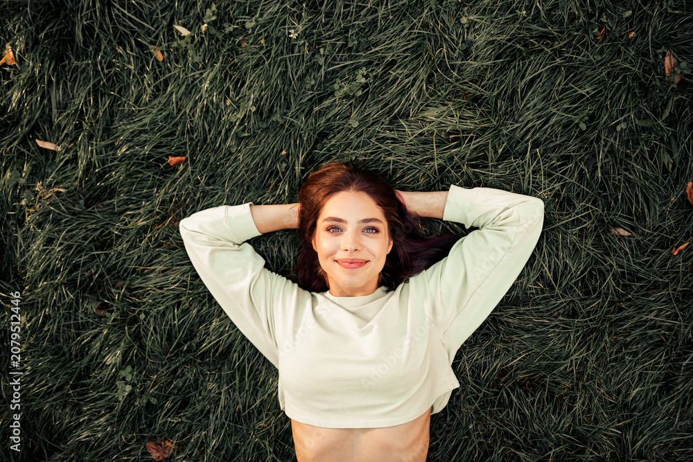 Fototapeta premium Young woman lying on green grass smiling and relaxing with hands behind her head in a casual cropped sweatshirt, enjoying outdoor nature and a peaceful moment