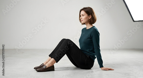 Side view of a beautiful young woman with short hair sitting on a studio floor, wearing a dark green sweater, black pants, and loafers, looking away in a minimalist setting.
