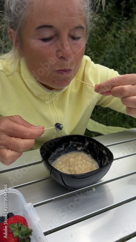Senior woman dipping fresh forest berries on toothpicks into sweet banana sauce at campsite.
