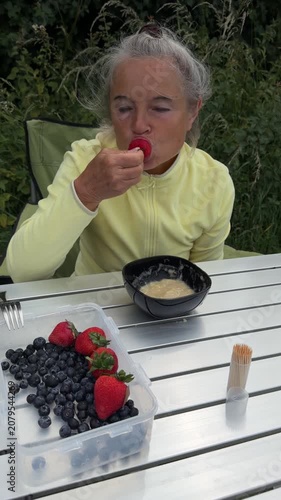 Senior woman enjoying sweet strawberry dessert with banana sauce at outdoor campsite table.