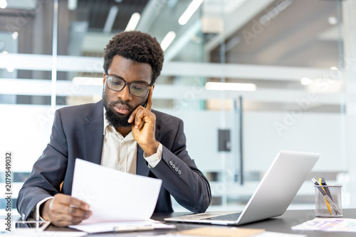 Fotografija Young businessman in eyeglasses sitting at office desk, concentrating on reading