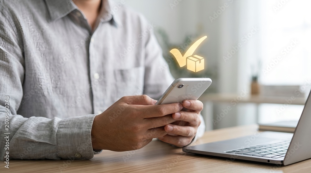 Fototapeta premium Smartphone displaying package delivery confirmation icon held by a man wearing a grey shirt sitting at a wooden desk