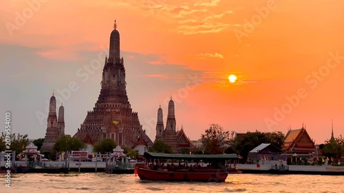 Wat Arun Ratchawararam in Bangkok, Thailand, with a bright golden orange sunset as the backdrop. The reflection of the temple on the water below creates a peaceful and elegant atmosphere