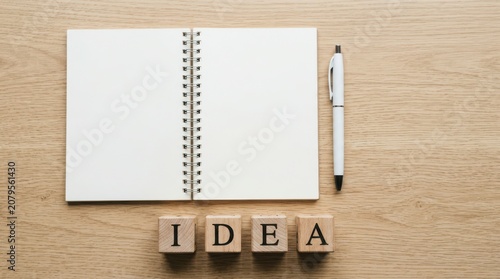 Open notebook with a pen and wooden blocks spelling the word idea sitting on a light brown wooden desk surface for creative work