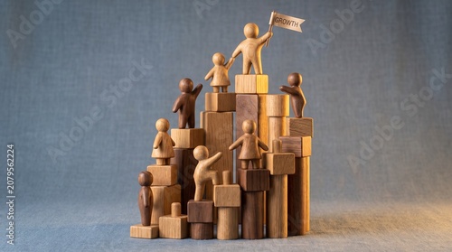 Wooden figurines climbing a staircase of blocks towards success with a leader holding a growth flag, symbolizing business development, teamwork, and achievement.