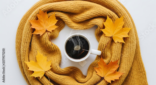 A steaming cup of hot black coffee placed in the center of a bright yellow knitted scarf surrounded by vibrant autumn maple leaves on white