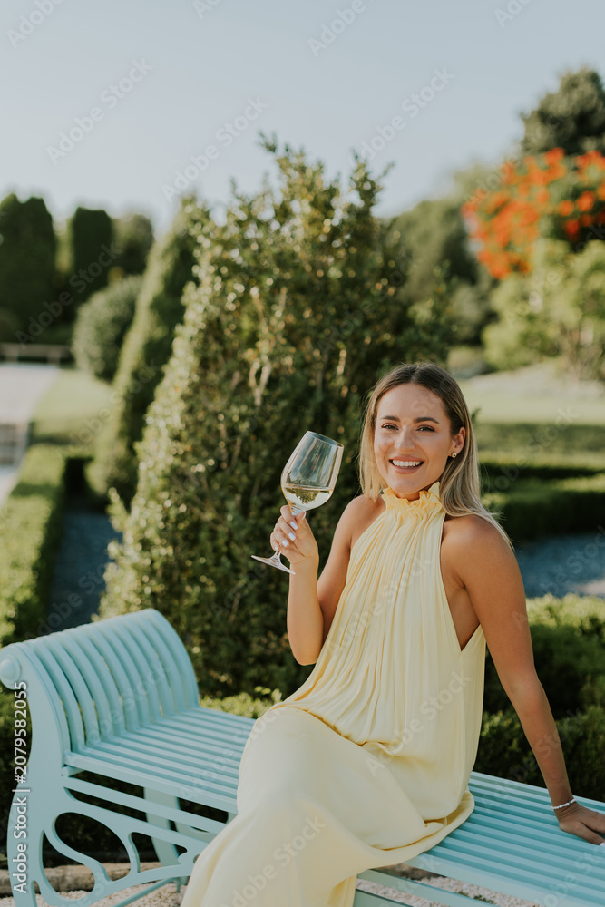 Fototapeta premium Woman sits on a blue bench in a garden holding a glass of wine during daytime