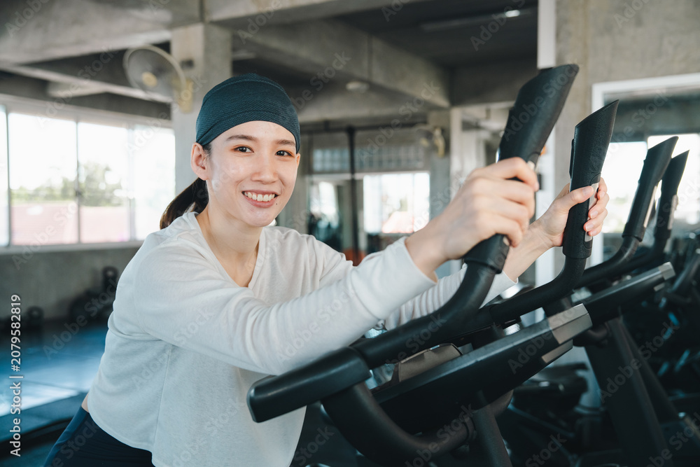 Fototapeta premium Woman gym exercise fitness elliptical machine smile indoor workout cardio, happy woman exercising elliptical machine indoor gym, wearing headband and white shirt, holding handle during cardio