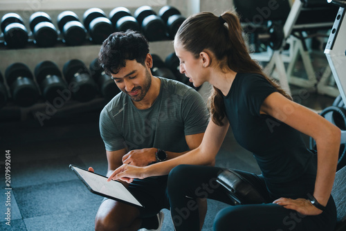 Personal trainer gym woman man clipboard workout consultation fitness coaching exercise, focused adult pair reviewing training plan near dumbbell rack on bench