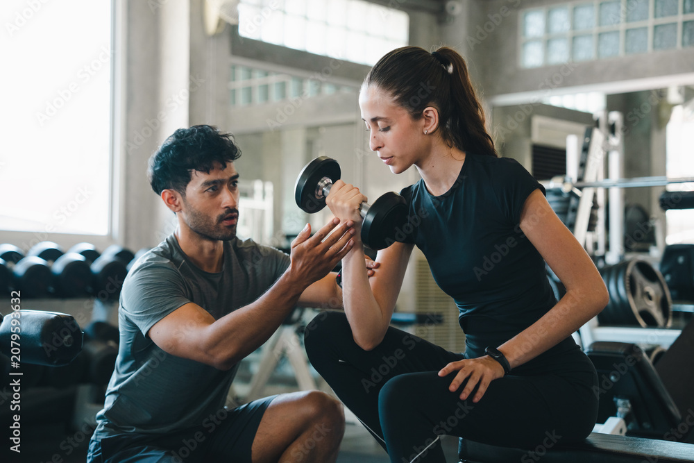 Fototapeta premium Fitness woman trainer dumbbell gym exercise coaching strength workout support, woman lifting dumbbell with male trainer guidance modern gym, focused expression during strength exercise