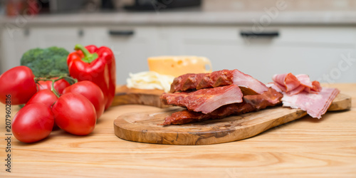Smoked pork ribs with ham, fresh vegetables and cheese on wooden cutting boards in a modern kitchen. Ingredients for homemade cooking and barbecue.