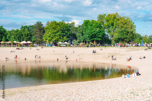 People relaxing around a small lake with a pebble beach in a green city park on a sunny spring day. Bundek lake, Zagreb, Croatia