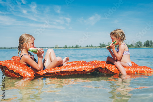 two young girls eating watermelon slices while relaxing on orange inflatable float mattress in lake water during sunny summer vacation representing childhood freedom friendship and outdoor leisure