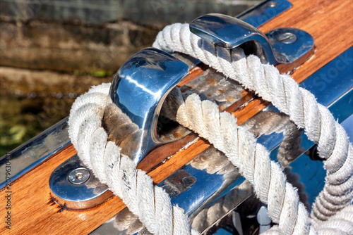A cleat is mounted on a wooden yacht with a rope tightly secured. The setting is at a harbor where boats are docked. This shows part of a sailing operation.