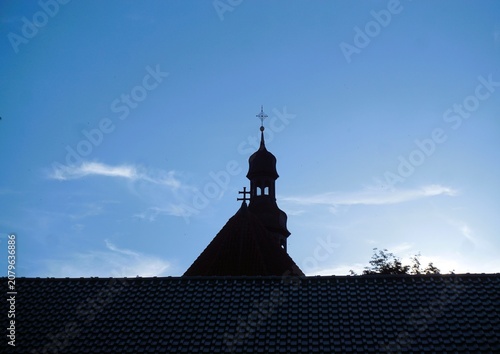 Schwarze Silhouette von Kirchturmpanorama hinter Hausdach und vor blauem Himmel mit weißen Wolken in Stadt bei Abendrot im Sommer 