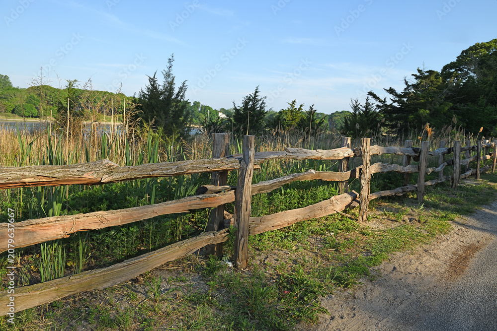 Fototapeta premium Picturesque landscape. Wooden fence along rural road in Poquott Village. Suffolk County, New York. United States