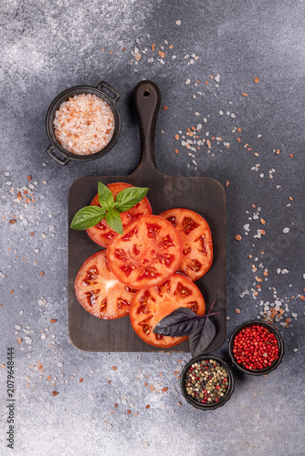 Sliced red tomatoes with basil, salt, and peppercorns on dark board