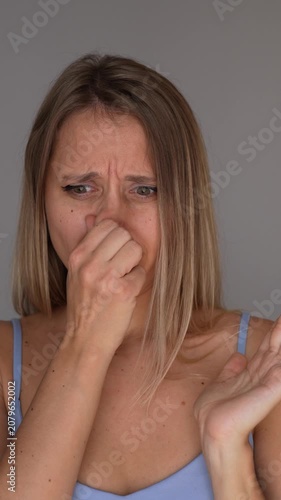 Young blonde woman notices a bad smell, closes her nose and waves her hand with a disgusted expression, showing rejection, discomfort and unpleasant odor in a hygiene concept on a grey background 