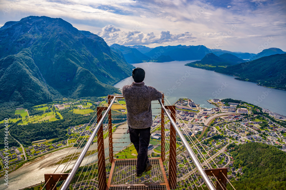 Obraz premium Hiker on Nesaksla mountain, enjoying a scenic view of Andalsnes, Norway, and the surrounding Rauma River, Romsdalsfjord and steep Romsdal peaks under dramatic sky.