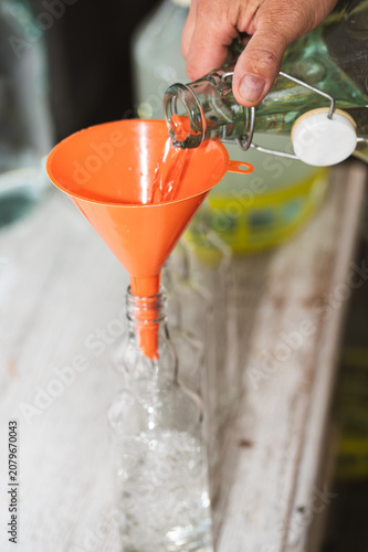 Photography Pouring clear alcohol from glass bottle into large demijohn carboy through orange plastic funnel, moonshining process
