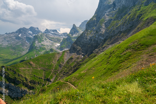 Lush green alpine slopes and rugged peaks near Engelberg, Switzerland, with distant snow patches and dramatic clouds on a clear summer day.