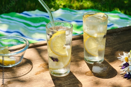 The process of preparing cold water with lemon and ice cubes in tall glasses outdoors in summer.