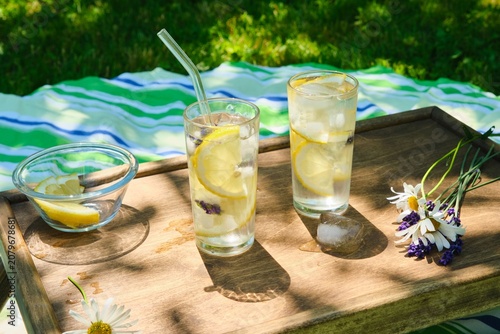 The process of preparing cold water with lemon and ice cubes in tall glasses outdoors in summer.