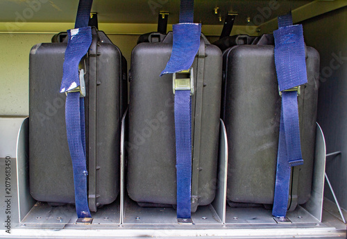 Fotografía Three heavy-duty black plastic protective equipment cases secured with blue tie-down straps in a storage compartment or vehicle shelf