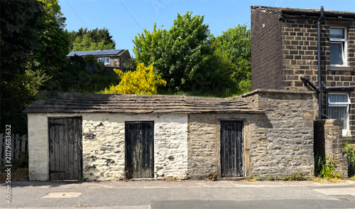 Old stone outhouses, with three wooden doors stands beside a stone building. Lush greenery and vibrant shrubs surround the structures on a summers day in Oakworth, Keighley, UK