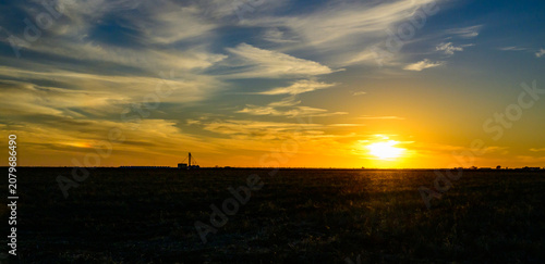 Amarillo, Texas. A serene prairie landscape at a vibrant sunset. The low sun illuminates the sky with brilliant orange hues above the silhouettes of rural buildings and agricultural infrastructure.
