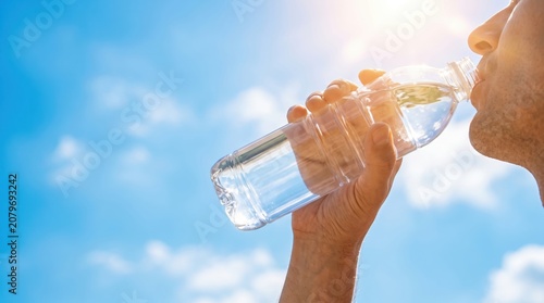 A person holds a plastic bottle and drinks water while standing outside. The sun shines bright in the sky, making the moment refreshing
