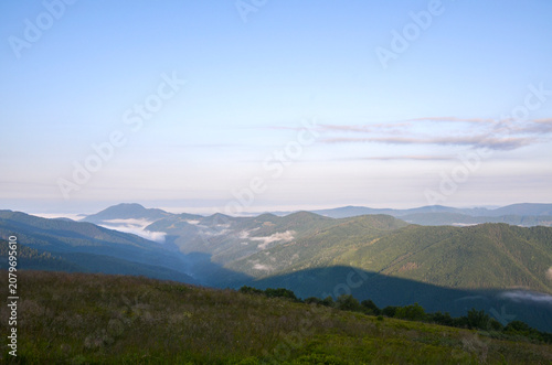 Soft morning light and low mist drift through rolling green mountains and deep valleys, with a grassy wildflower meadow in the foreground. Carpathian Mountains, Ukraine
