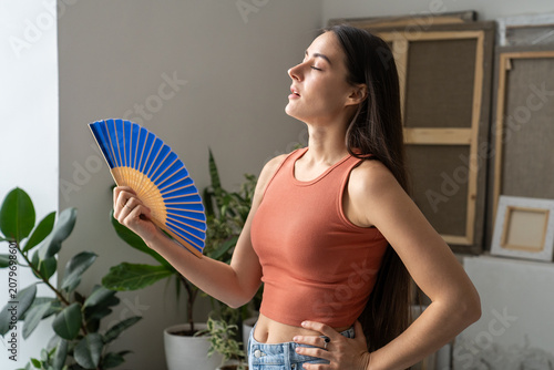 Foto Woman suffering from overheating at home without air conditioner, stands beside window waving hand paper fan for cooling herself