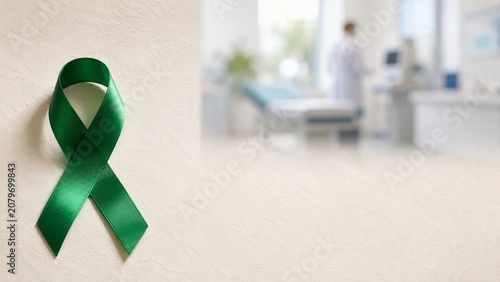 Green awareness ribbon on a textured table with copy space, against a blurred clinic background with a doctor, symbolizing medical support for liver cancer, lymphoma, or mental health campaign.
