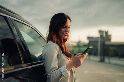 Woman standing by her car, smiling while texting on a smartphone, enjoying sunset light. Connecting on the go