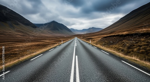 A straight asphalt road stretches through barren mountains under dramatic cloudy skies. Brown hills and rocky terrain line the highway with yellow center markings. Perfect for travel and adventure.