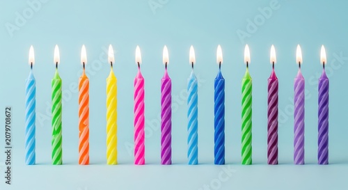 Colorful birthday candles with lit flames against a soft blue background. The candles feature spiral stripes in vibrant colors including blue, green, orange, yellow, pink, and purple.