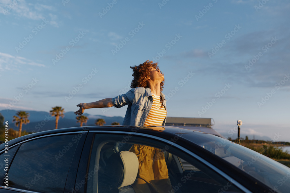 Obraz premium Woman with curly hair leans out of a sunny car window, enjoying coastal breeze and carefree lifestyle, palm trees and ocean visible during golden hour against a clear sky, distant shoreline, warm