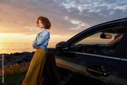 Quadro su tela Cheerful woman by a sleek car at sunset, enjoying a relaxed lifestyle moment, de