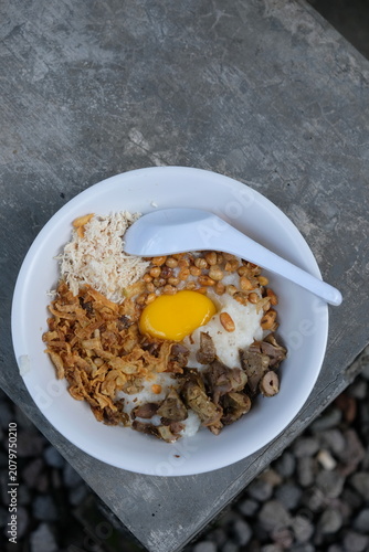 Bubur ayam. Chicken porridge topped with shredded chicken, fried soybeans, raw egg yolk, fried shallots, and minced chicken liver and gizzard. Served in a white ceramic bowl. Indonesian food.