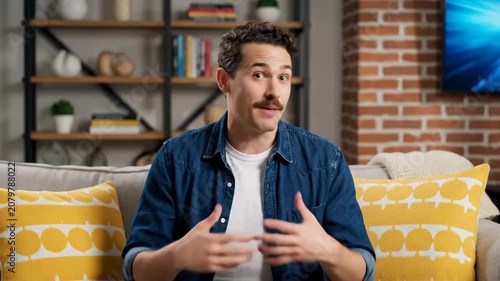 Young man with a mustache talking and gesturing on a sofa in a modern living room.