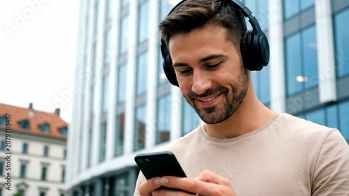 Smiling young man in headphones using smartphone outdoors, enjoying music and communication in the city.
