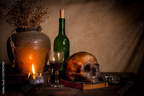 Human skull and candle with on wooden floor and old dirty wall background, and vase flower,and glass,and bottle of wine, still life concep
