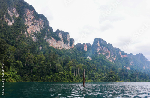 Limestone cliffs and dense tropical forest rise above a calm green lake under a soft cloudy sky.